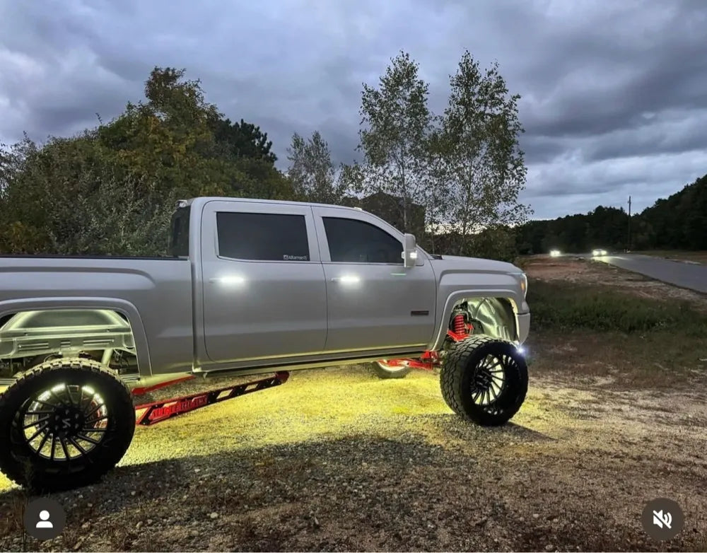 Lifted silver pickup truck with large custom wheels and underglow lighting parked outdoors at dusk.