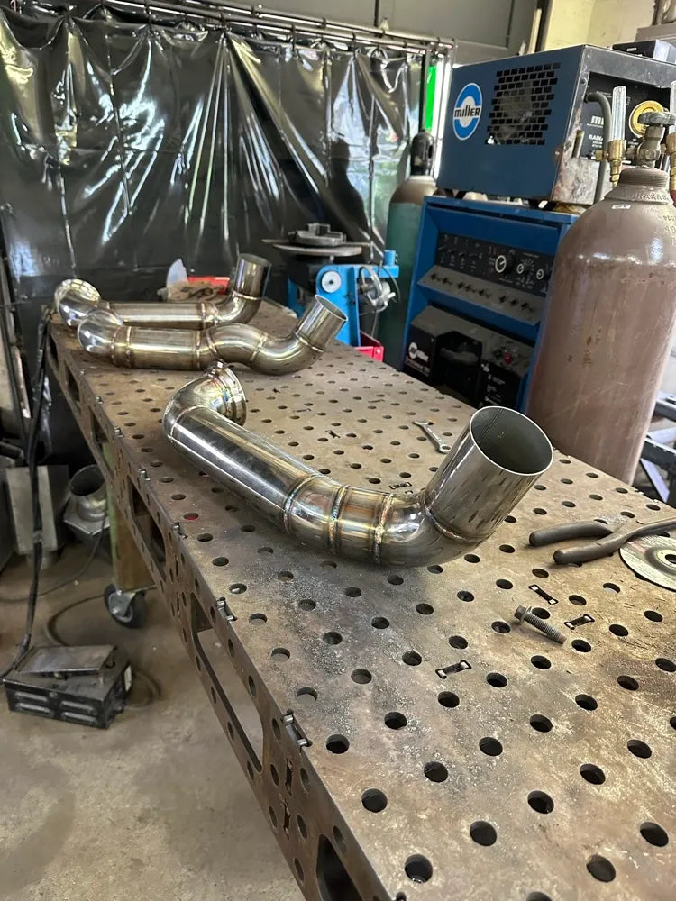 Welded stainless steel exhaust pipes on a perforated metal workbench in a fabrication shop
