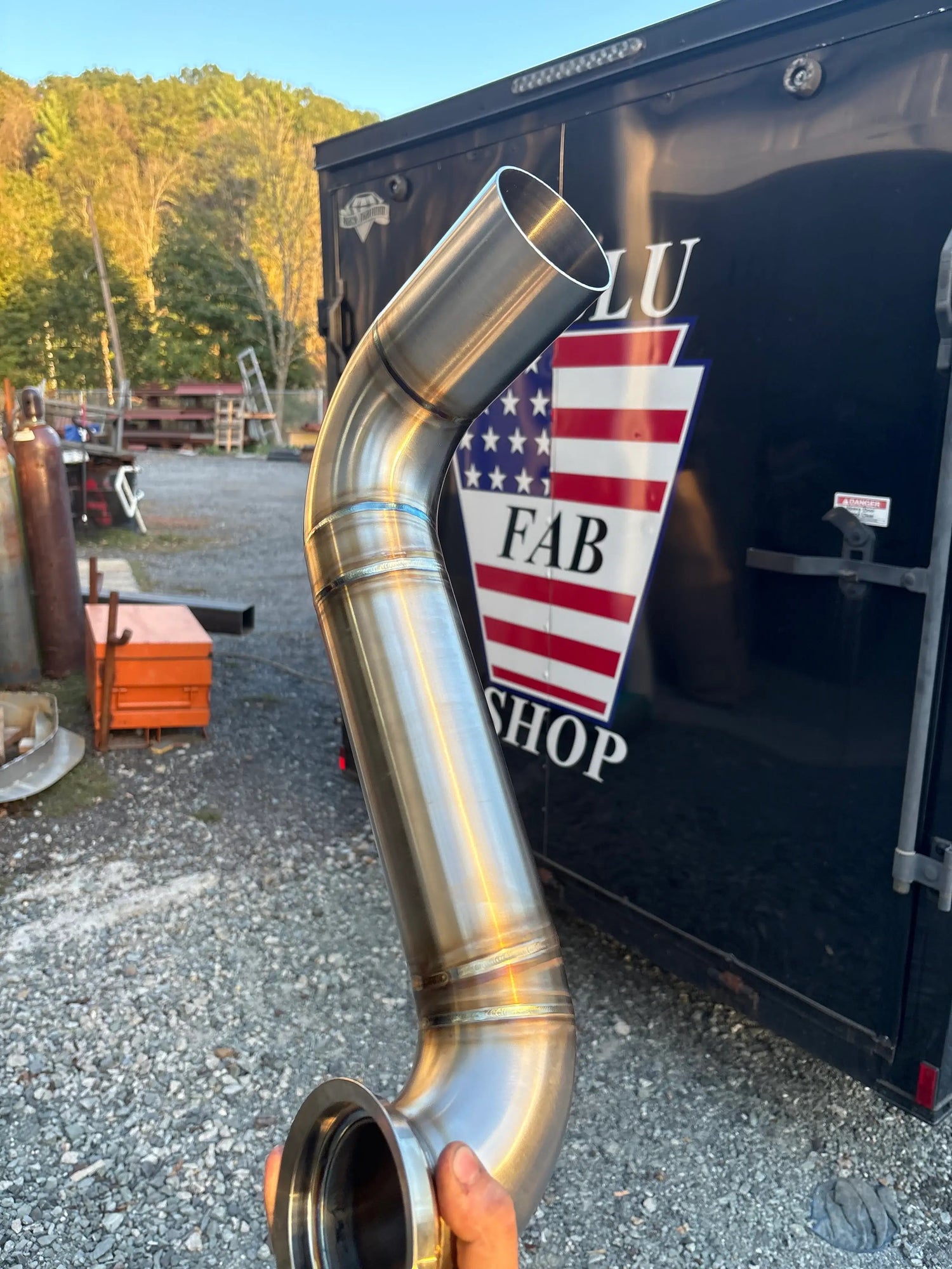 Custom stainless steel exhaust pipe with polished welds, held outdoors near a workshop trailer