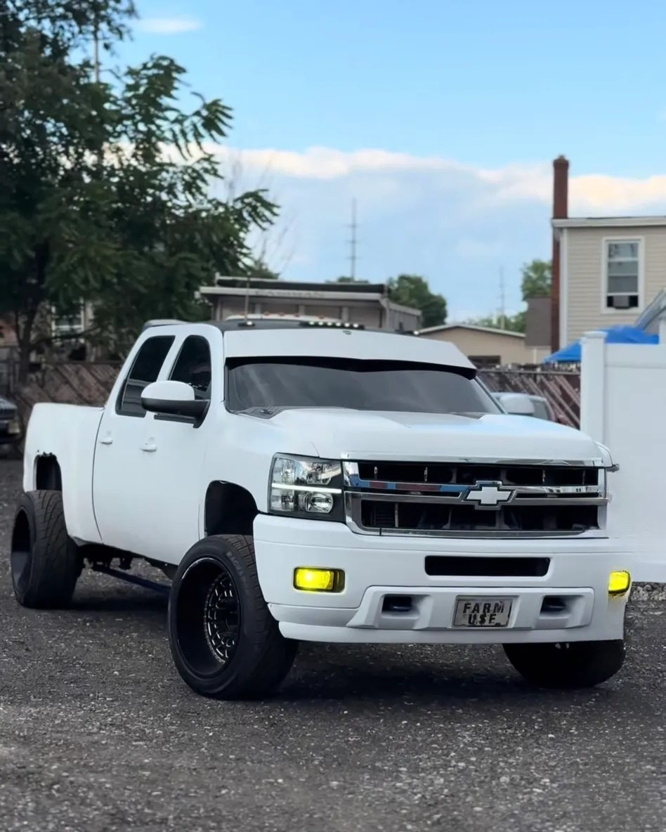 White Chevrolet pickup truck with custom wheels and yellow fog lights on gravel driveway