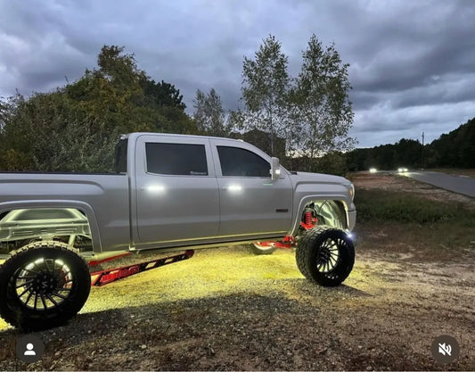 Lifted silver pickup truck with oversized wheels and glowing undercarriage lights at dusk