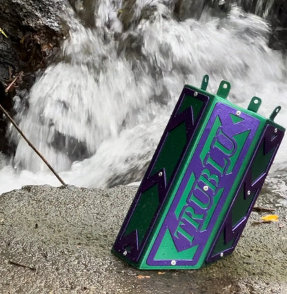 Purple and green metal trap box with TRUBLU text, placed outdoors near a flowing stream