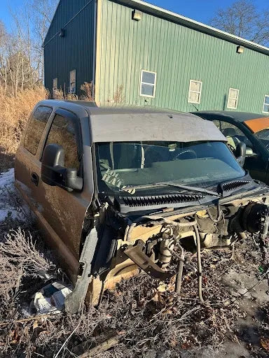 Abandoned tan SUV with missing front end parts in overgrown lot next to green metal building