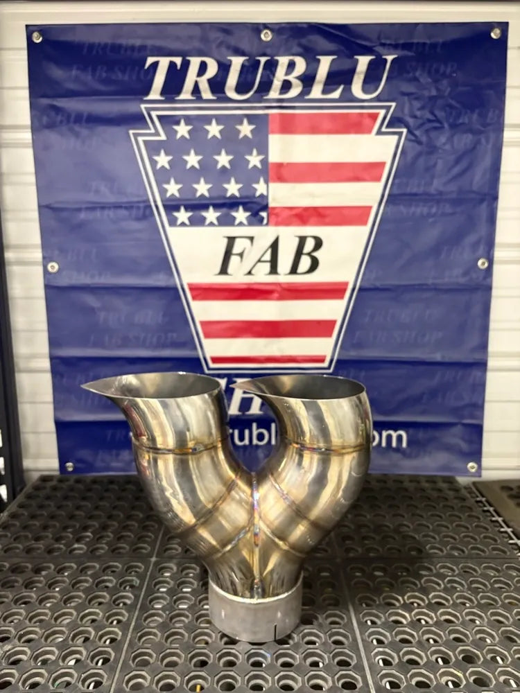 Polished stainless steel custom exhaust Y-pipe on metal workbench in a workshop.