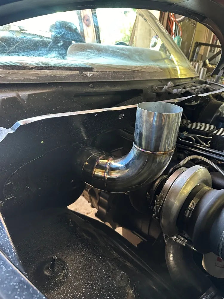 Polished custom turbo downpipe installed in a car engine bay, showing detailed welds.