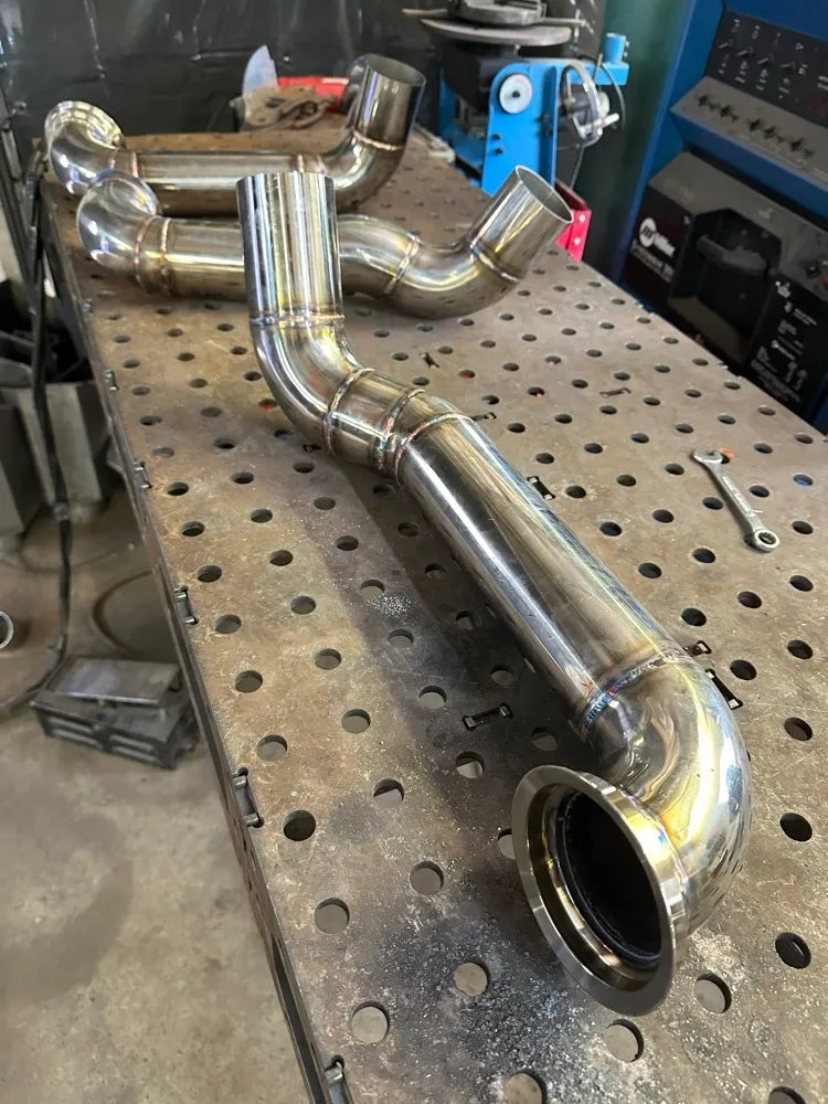 Welded stainless steel turbo downpipes on a metal workbench in a fabrication shop