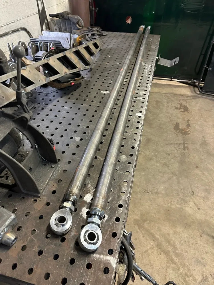 Two metal suspension link bars with rod ends on a perforated welding table in a workshop