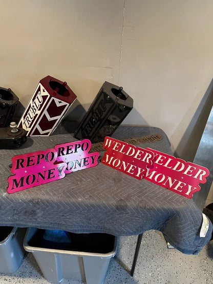 Custom metal signs reading 'REPO MONEY' and 'WELDER MONEY' on display table, fabricated parts in background.