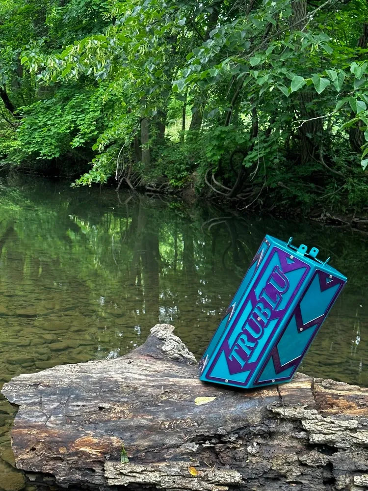 Colorful metal box with TRUBLU text on log by forest stream, nature background